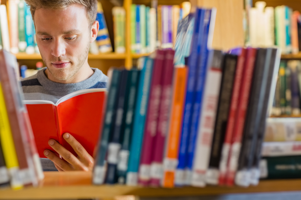 Young male student reading a book amid bookshelves in the college library